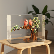 display of two pink lego birds on a branch inside a clear glass case on a wooden table.