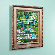 A framed lego art print depicting a vibrant green garden scene with a blue arched bridge over water lilies, mounted with a brown acrylic frame.