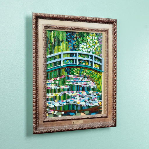 A framed lego art print depicting a vibrant green garden scene with a blue arched bridge over water lilies, mounted with a brown acrylic frame.